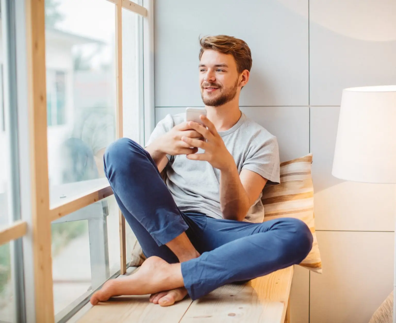Young man enjoying at home. Sitting on window wooden seat and using smart phone.